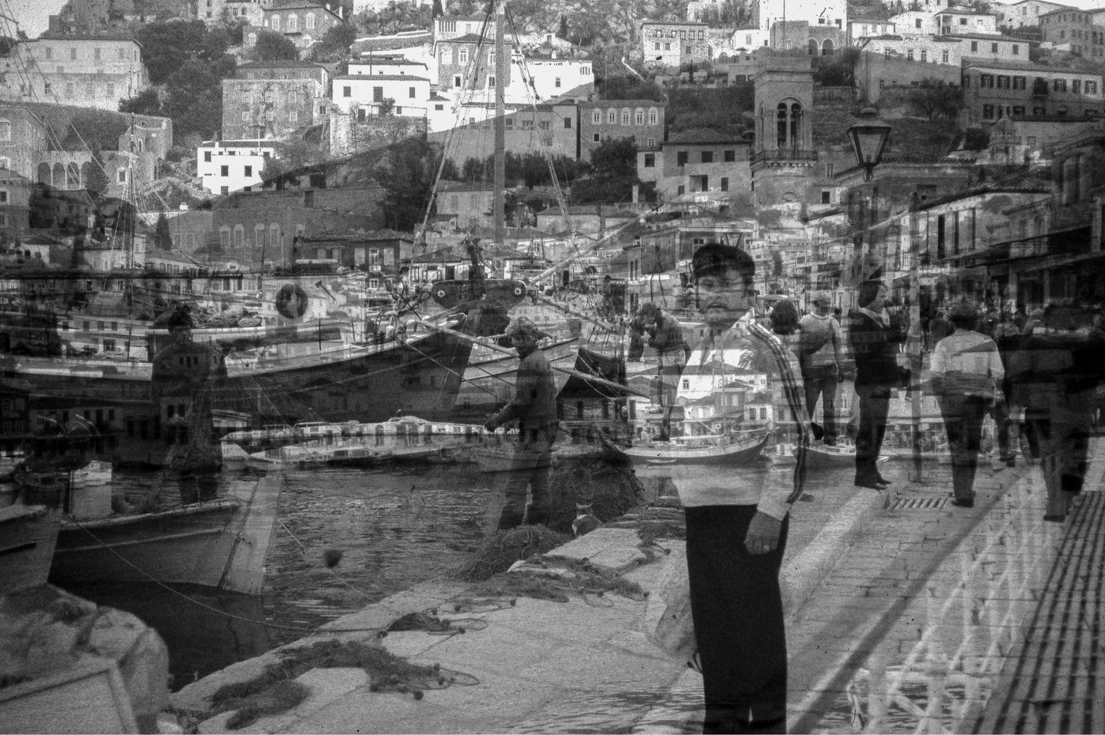 Black-and-white overlap of a harbor town; the artist’s grandfather stands on the quay, boats and hillside houses passing through him.