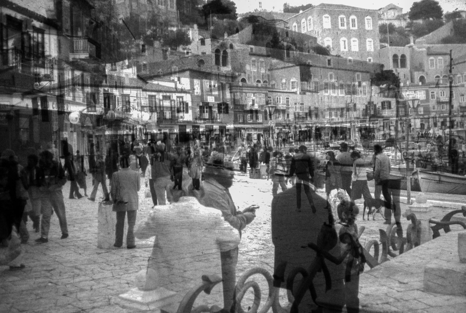 Black-and-white overlap of a busy harbor square, a man in a hat at center amid ghosted crowds and boats.