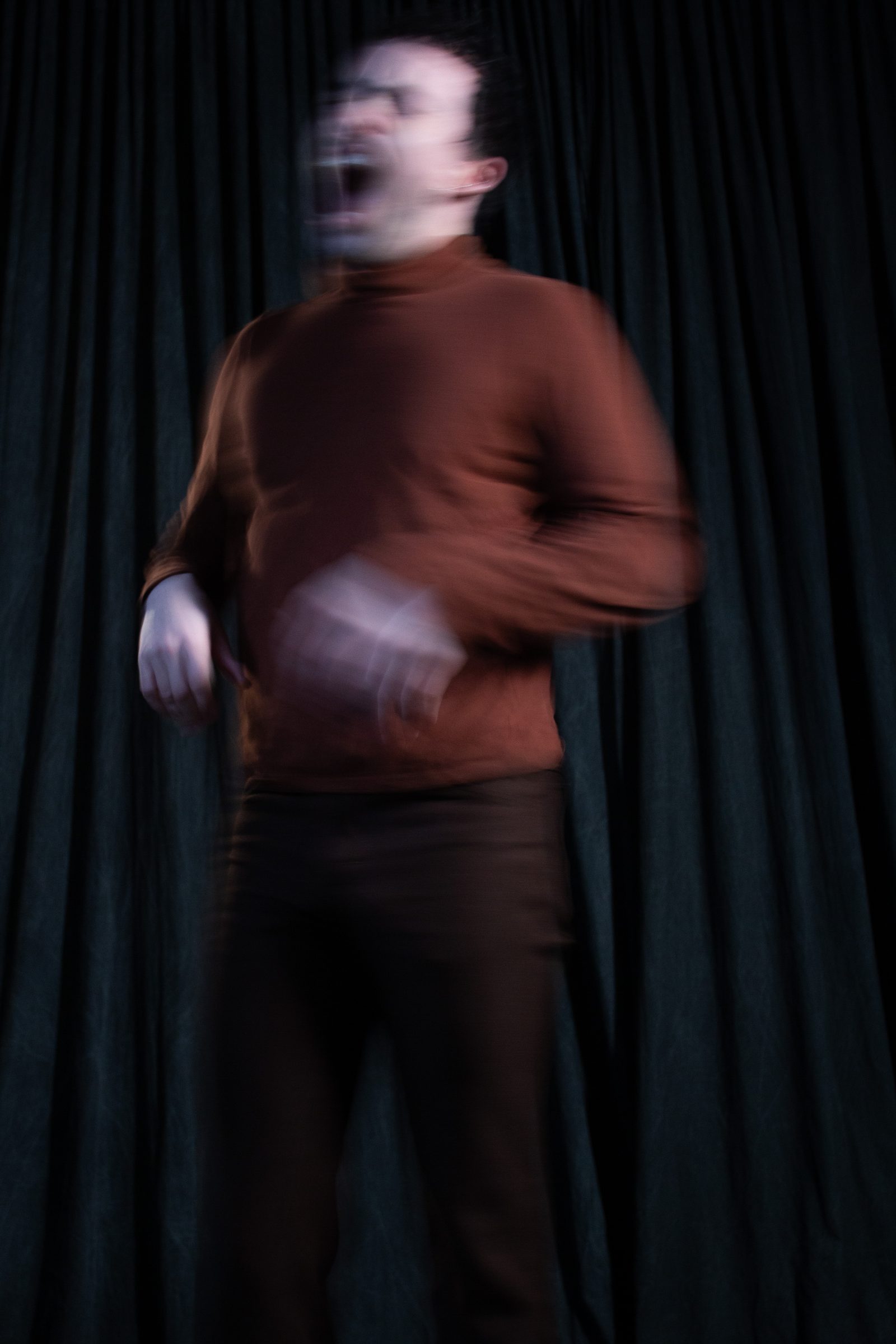 The artist in a rust turtleneck, mouth open mid-shout, face and hand blurred by motion against a dark curtain.