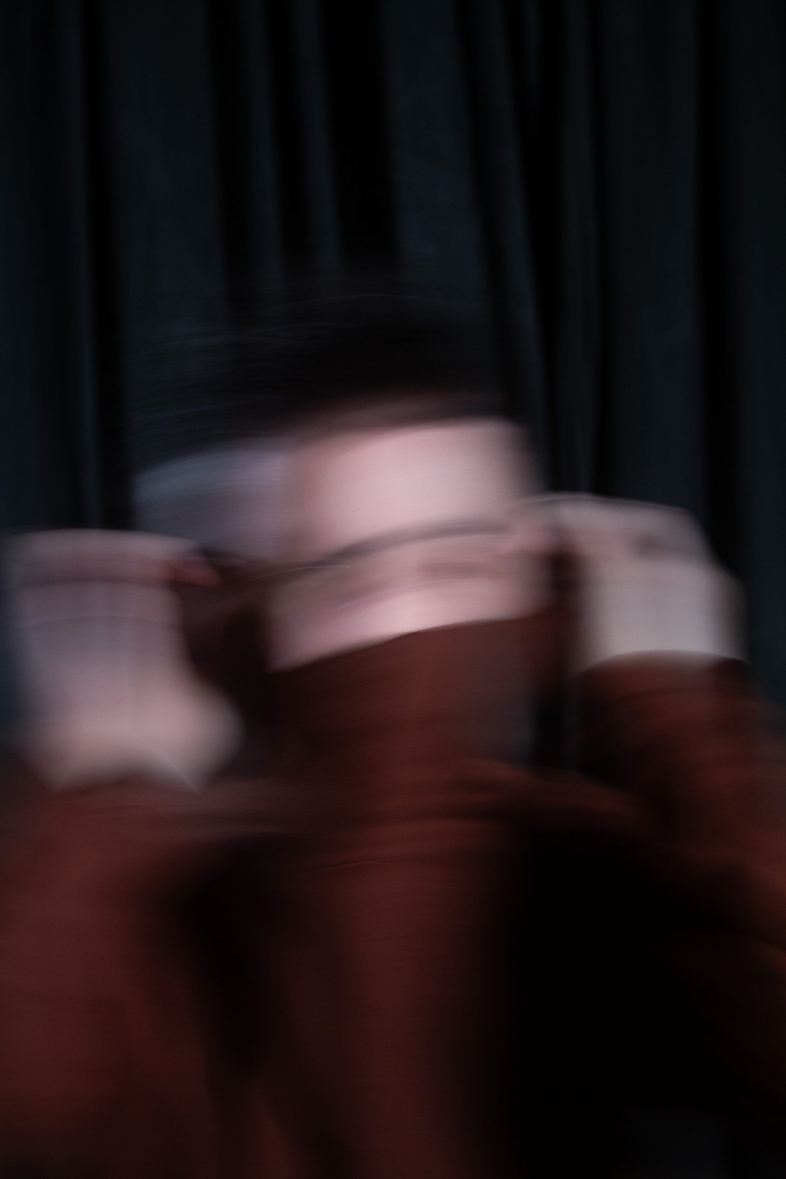 The artist’s face hidden behind raised hands, everything smeared by motion in rust and dark teal.