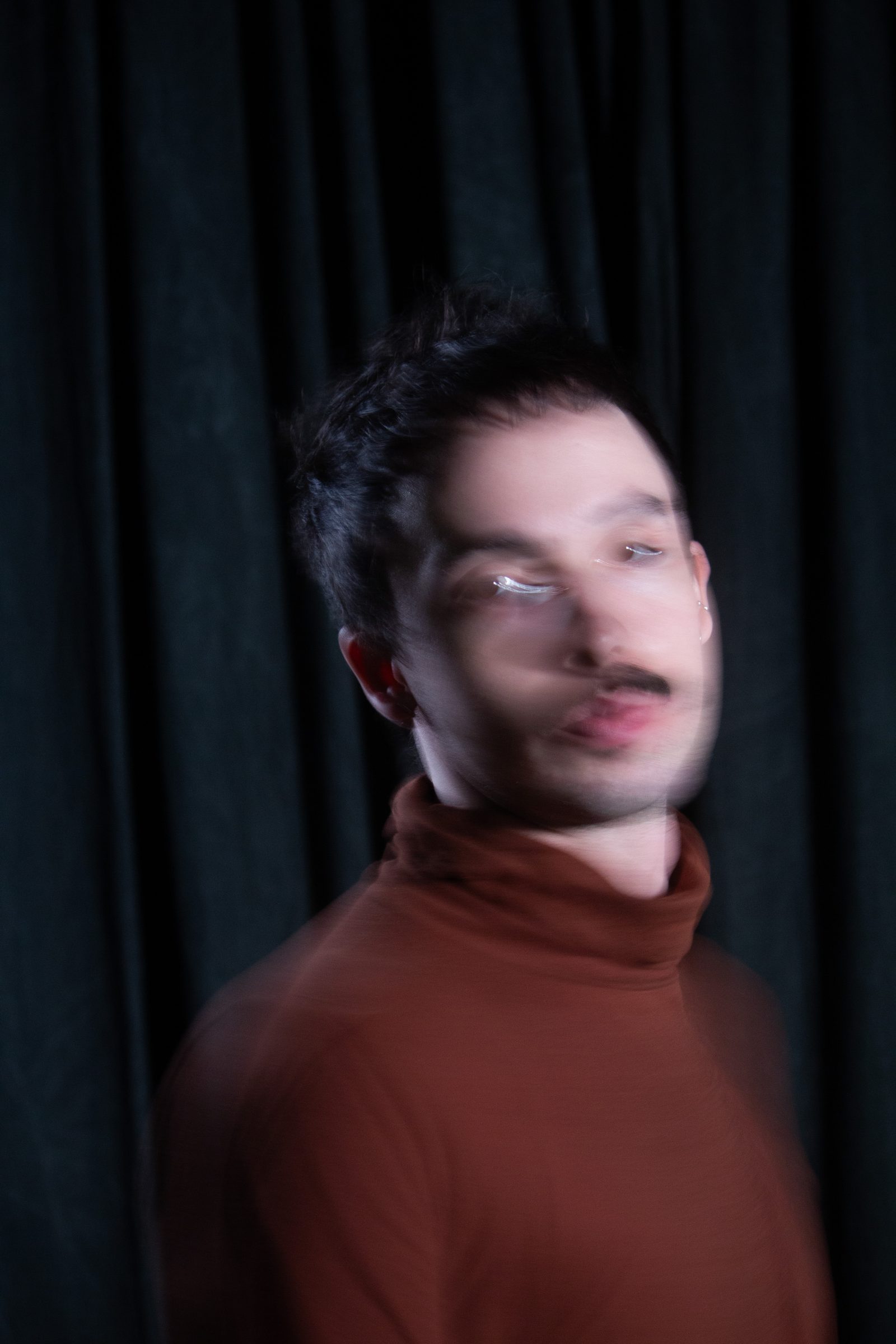The artist turning, face stretched and doubled by motion blur, in a rust turtleneck against a dark curtain.