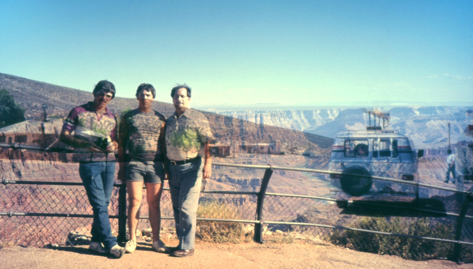 Three men at a canyon railing under clear blue sky, a parked van drifting translucent beside them.