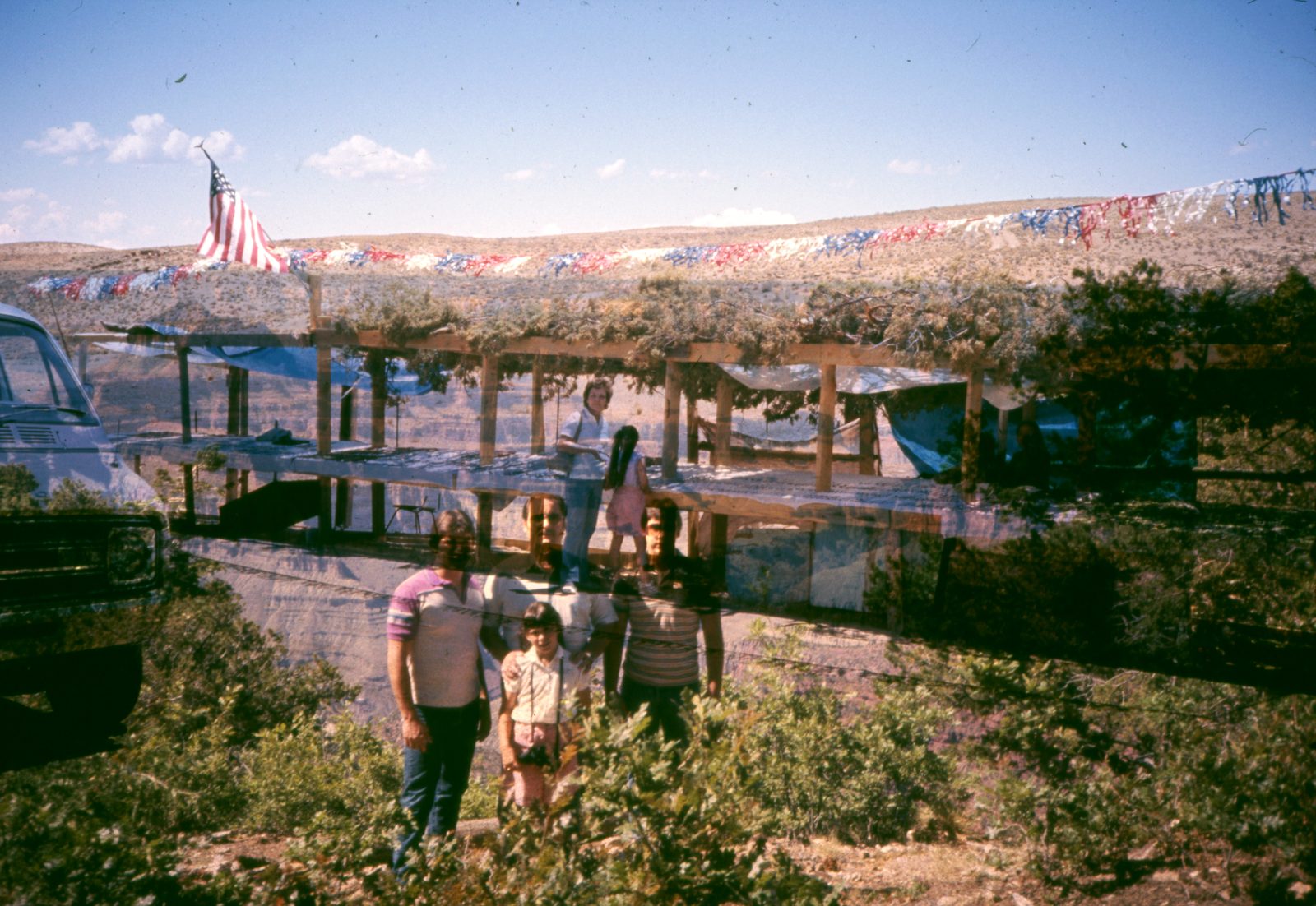 Family in front of a wooden shelter strung with flags, blending into a scrubby hillside.