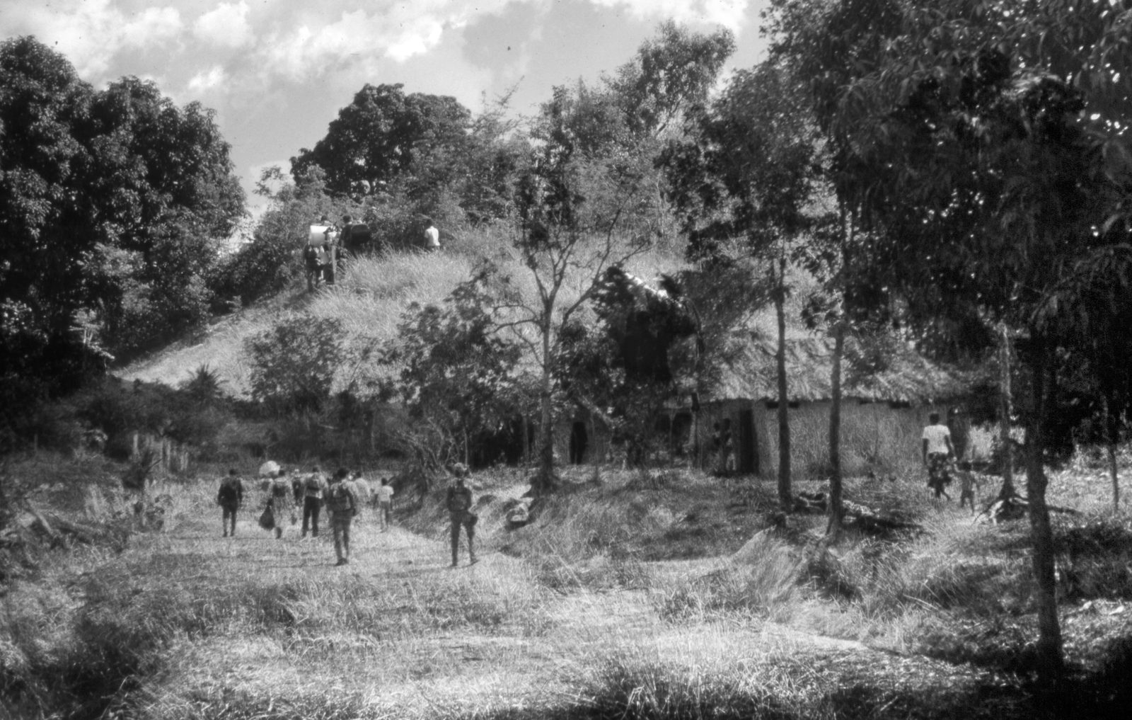Black-and-white view of figures walking a grassy path toward thatched huts and a hill, framed by dense trees.